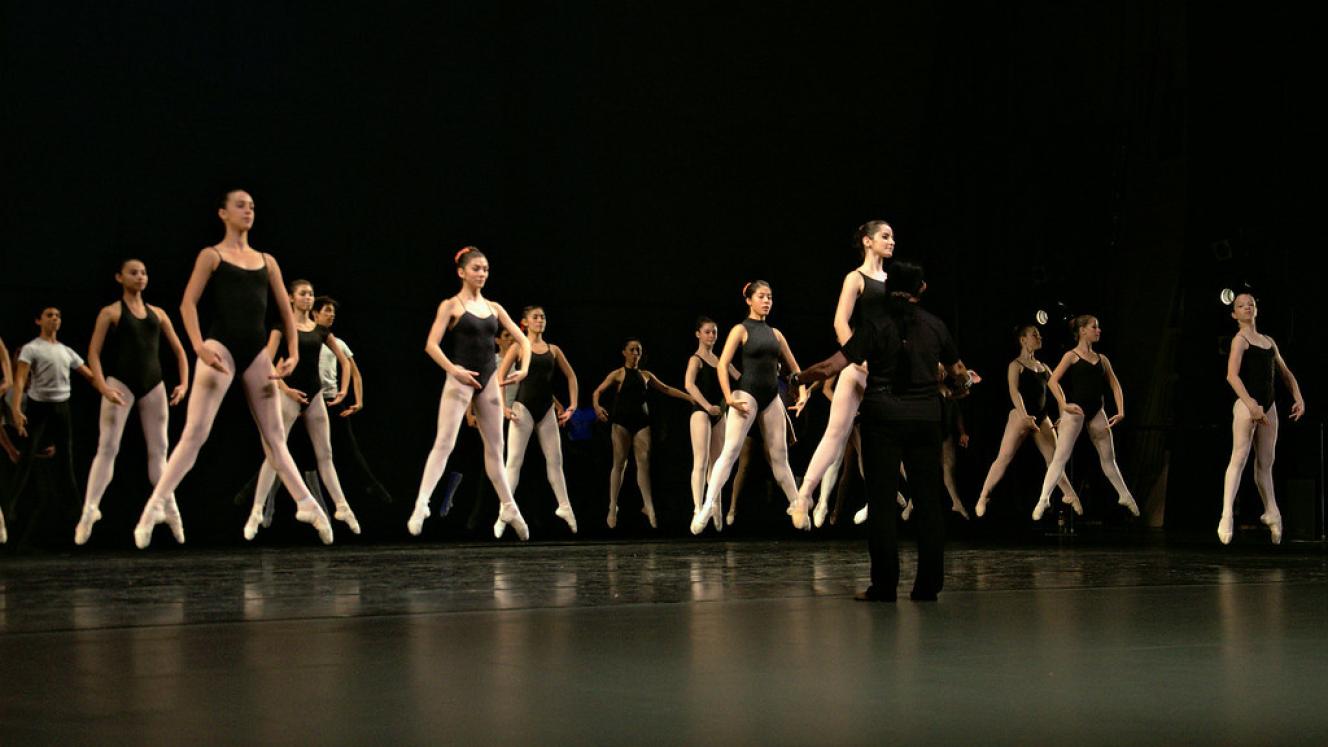 Rows of ballet dancers in black leotards and pink tights rehearse jumps together on a dark stage.